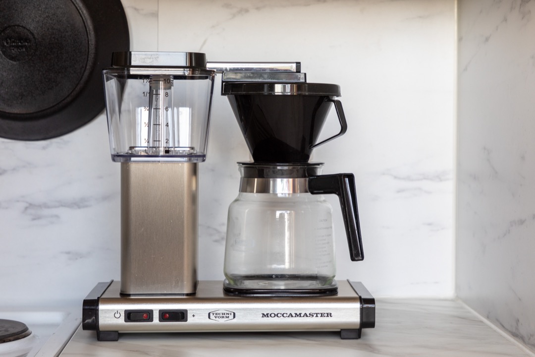 An automatic pour-over coffee machine brewing through a filter cone into a glass carafe on a kitchen counter