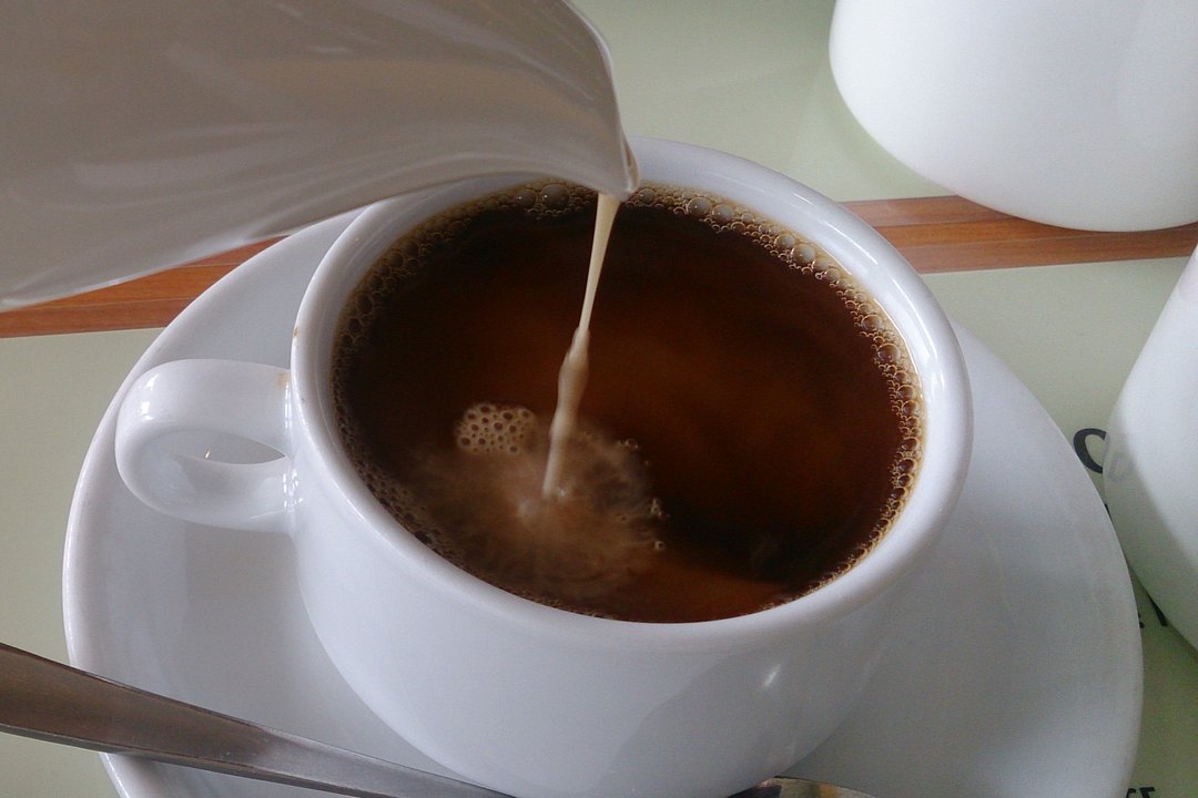 Cream being poured from a jug into a white cup of black coffee