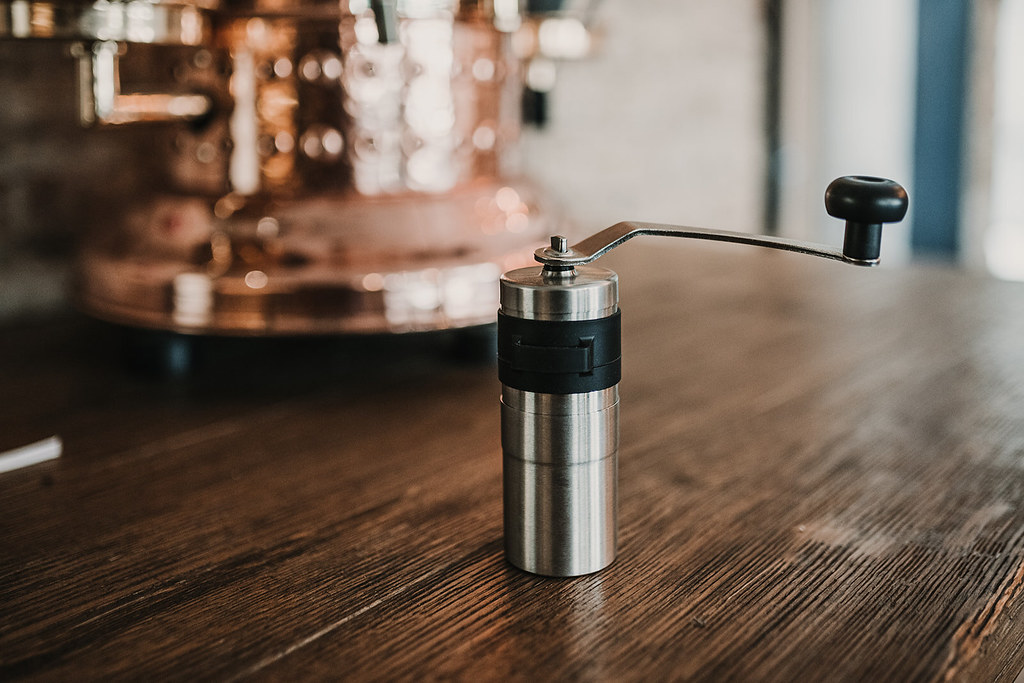 A stainless steel manual hand coffee grinder with a folding crank on a wooden counter beside a copper espresso machine