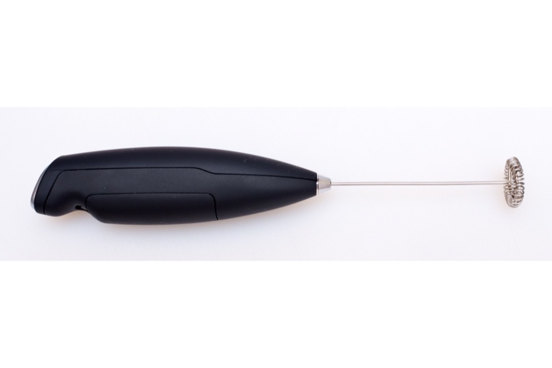 A handheld electric milk frother whisking a mug of frothed milk beside a cup of coffee