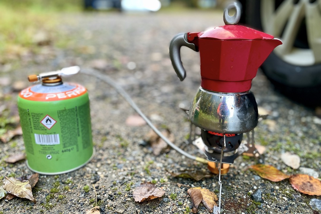 A red and stainless steel moka pot brewing on a portable gas burner outdoors