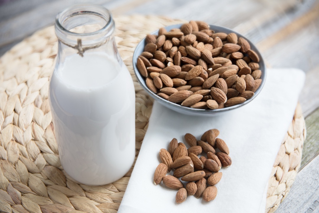 A glass bottle of almond milk standing beside a bowl of whole almonds on a light surface