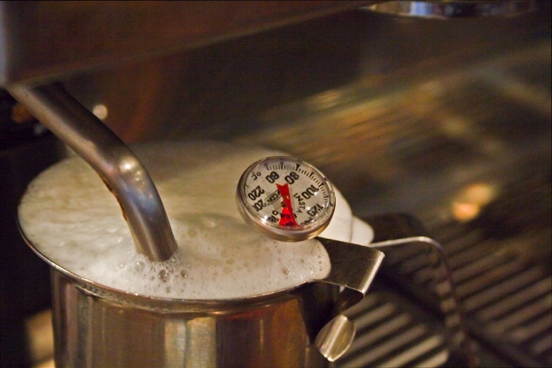 Steam wand texturing milk in a stainless pitcher with a clip-on thermometer reading the temperature