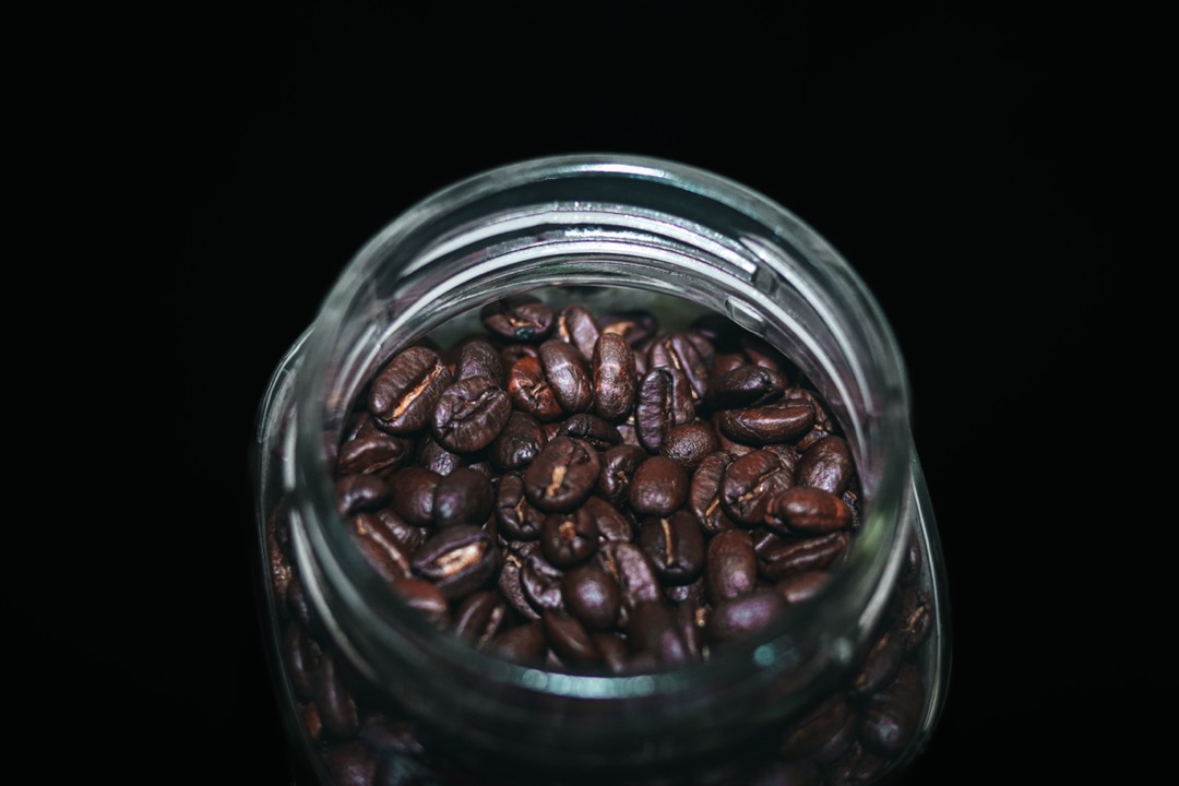 Roasted coffee beans inside a clear glass storage jar photographed from above against a dark background