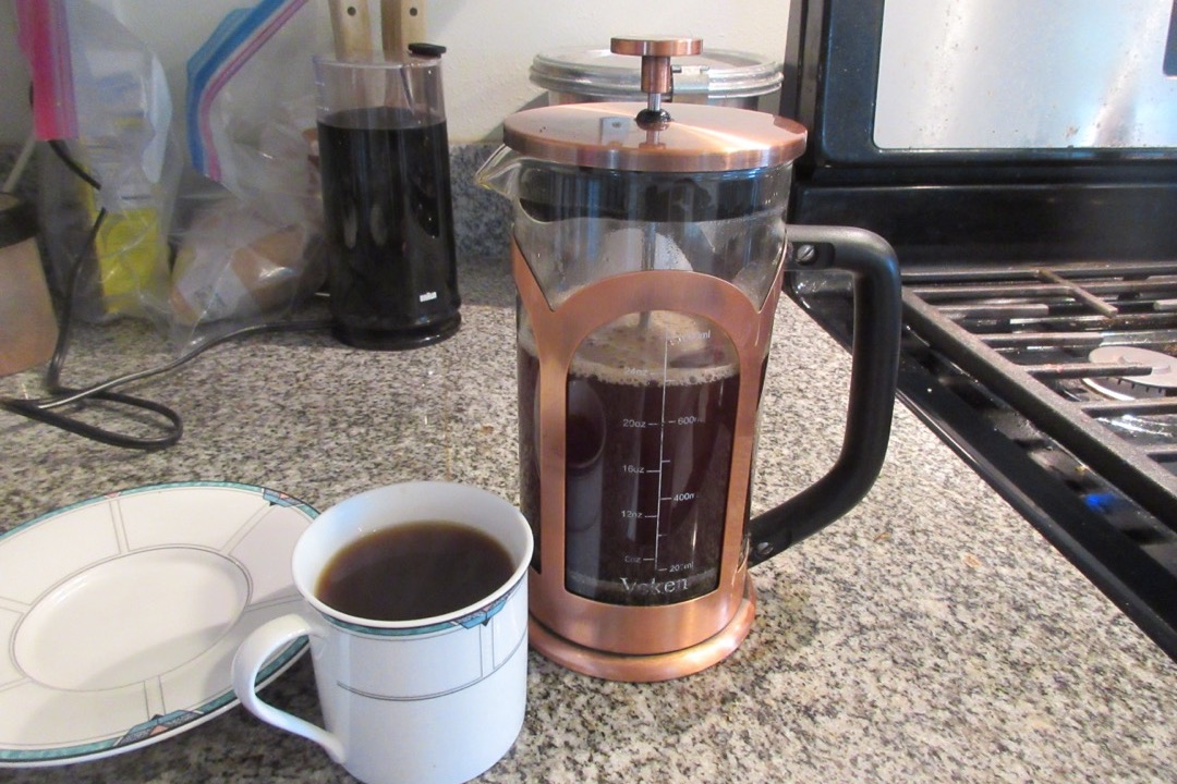 A glass French press full of freshly brewed coffee with the plunger pressed down, beside a mug of black coffee