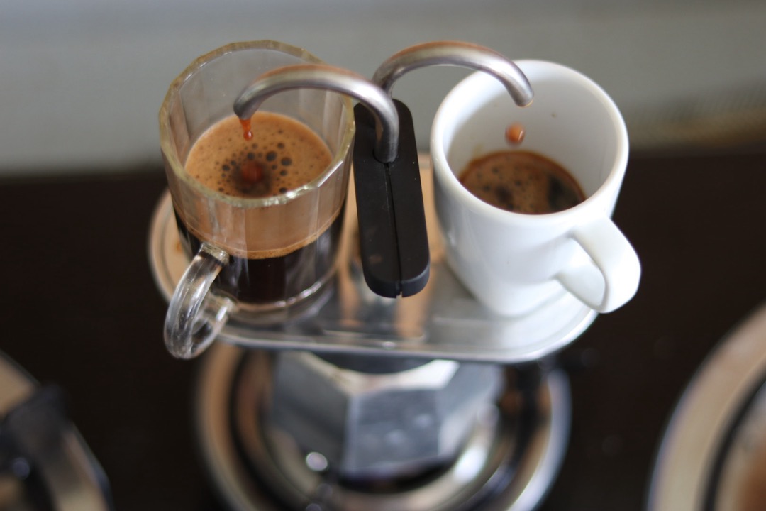 A stovetop moka pot brewing dark coffee directly into a glass and a white cup